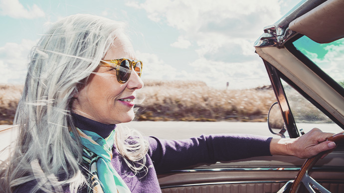 Woman from the baby boomer generation driving a convertible on a sunny fall day with her long, silver gray hair blowing in the wind.