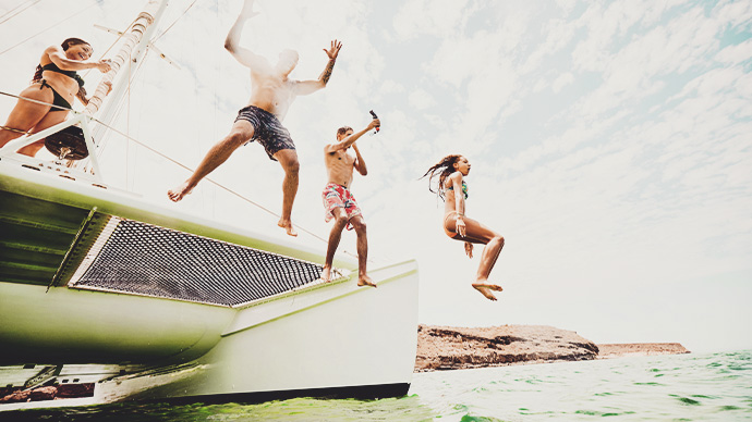 Wide shot of mother filming family jumping into tropical ocean from deck of sailboat while on vacation