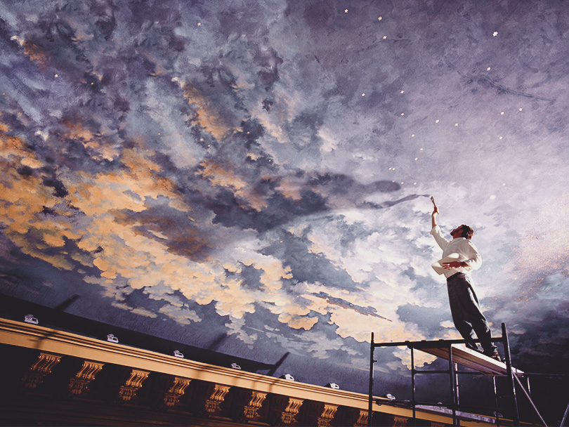 looking up on a man painting a cloudy and starry night sky mural on the ceiling