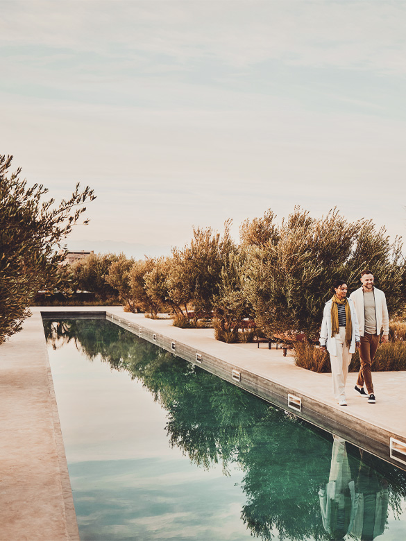 Wide shot of smiling couple on vacation walking along edge of pool surrounded by olive trees at boutique luxury resort in Morocco at sunrise